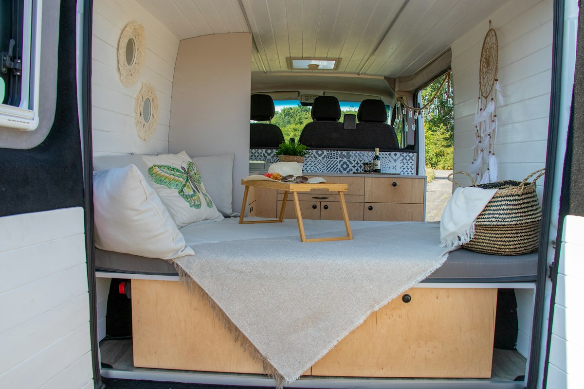 Interior de una furgoneta camper con una cama sobre cajones de madera y una mesita plegable encima.