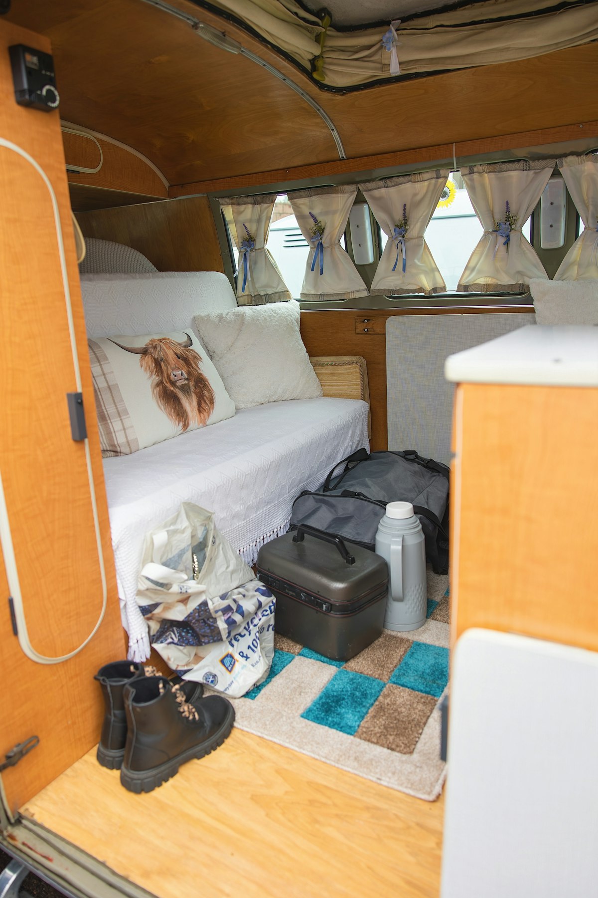 Interior de una furgoneta camper con cama, bolsas, botas y una nevera portátil en el suelo.