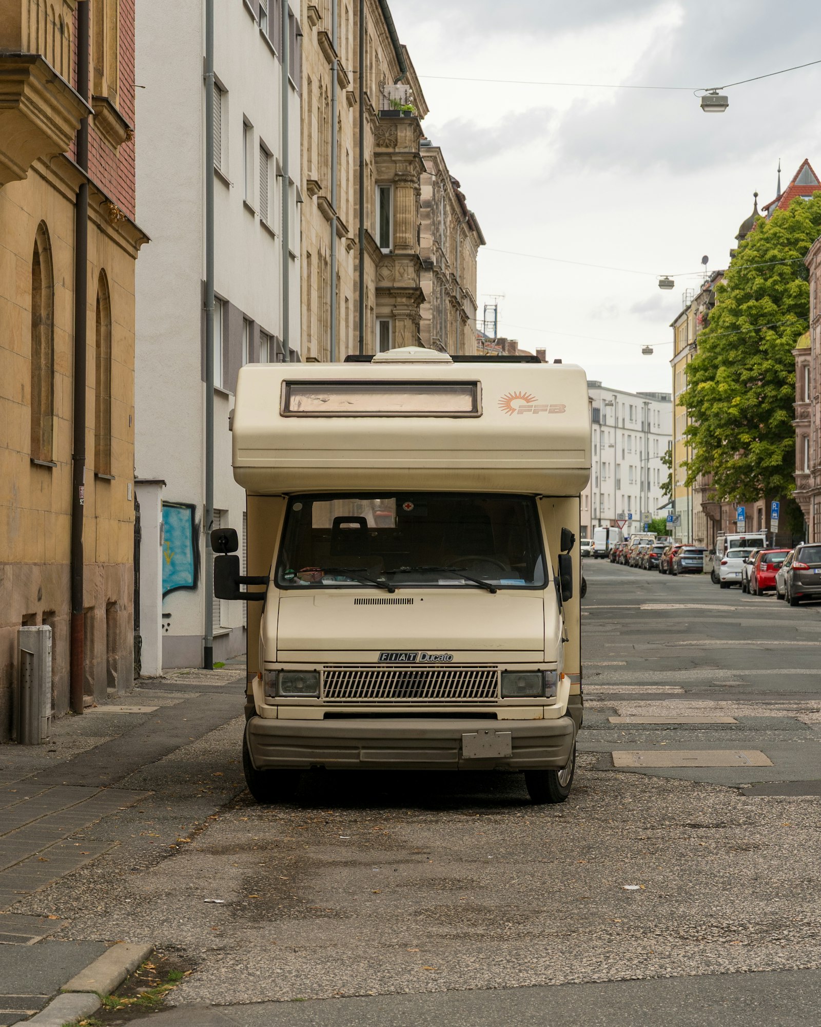 Autocaravana Fiat Ducato antigua de color crema aparcada en una calle estrecha entre edificios.